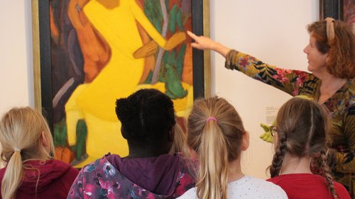 Kinder in Rückenansicht, die ein Gemälde ansehen, auf das eine Kunstvermittlerin zeigt.
